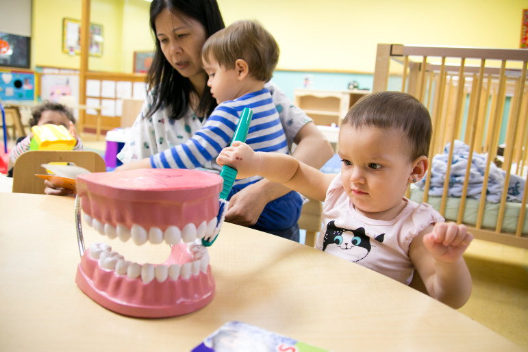 16-month-old Livia practices brushing teeth while 21-month-old Gunnar sits in Infant Care Aid Jian Liang's lap reading a book at the Magical Years Early Childhood Center, part of the Lutheran Family Health Centers.